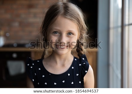 Headshot portrait of happy joyful little girl of primary school age with funny pigtails posing at home indoors alone looking at camera with naughty beautiful smile, making video call, profile picture