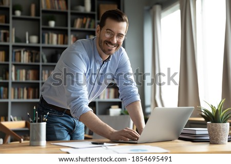 In modern office good-looking businessman young entrepreneur successful business owner standing leaned over workplace desk smile look at camera feels confident. Freelancer or teacher portrait concept Royalty-Free Stock Photo #1854695491