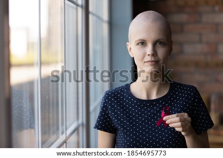 Symbol of hope. Profile picture portrait of young hairless woman diagnosed with oncology standing by window looking at camera holding scarlet pink ribbon meaning breast cancer awareness, copy space