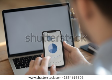 Businessman holding smart phone with financial data blue graphs charts on screen, laptop empty monitor with mock up, close up view over man shoulder. Synchronize information, new app, software concept