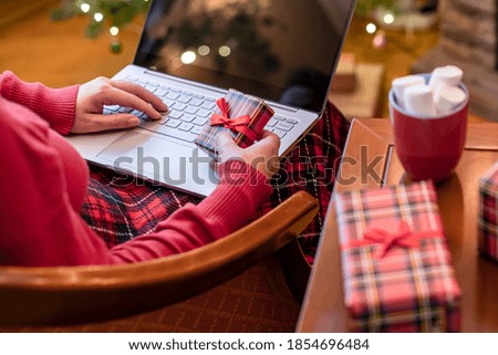 Christmas. Woman using laptop for searching gift ideas sitting at table with ready gift boxes and cup of cocoa and marshmallows near fireplace and christmas tree. Concept