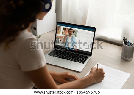 Back view focused young woman in headphones looking at computer screen, holding video call online private lesson with male teacher native speaker, learning foreign language distantly at home. Royalty-Free Stock Photo #1854696706