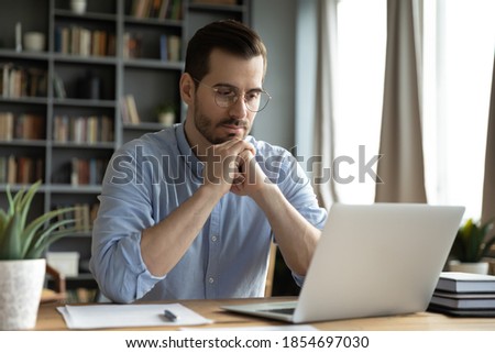 Young businessman sit at home office desk looks at laptop read media news online, learn new e app, analyzing project, stuck with challenge business task, thinks over problem, search solution concept Royalty-Free Stock Photo #1854697030
