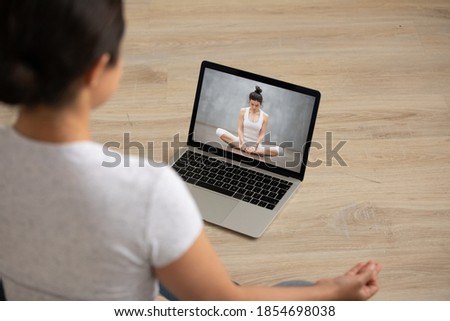 Focus on laptop screen with professional female yoga instructor sitting in lotus position, giving online master class. Young mixed race woman repeating exercises from video, enjoying training at home. Royalty-Free Stock Photo #1854698038