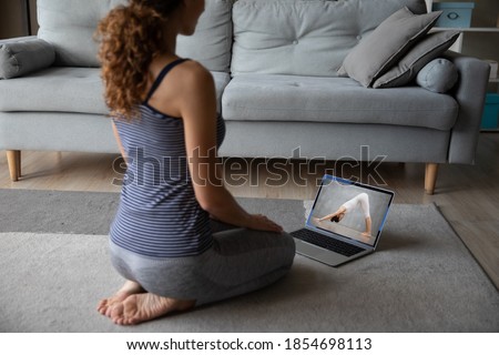 Back rear view happy young woman in activewear sitting on floor carpet, watching online yoga master class or educational webinar with professional female trainer standing in downward facing dog pose. Royalty-Free Stock Photo #1854698113