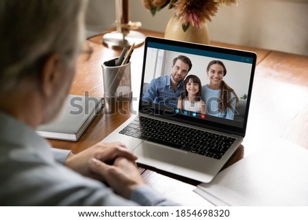 Rear shoulder view old senior middle aged man holding video call with happy young married couple grown kids and small granddaughter, enjoying pleasant conversation, distant communication concept. Royalty-Free Stock Photo #1854698320
