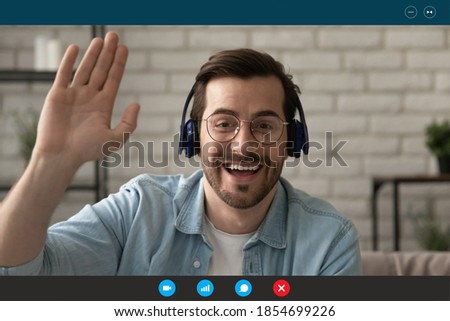 Head shot emotional happy 30s man in eyeglasses and wireless headphones, looking at camera, making hello gesture, greeting friends starting online video call, computer application monitor view. Royalty-Free Stock Photo #1854699226