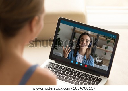 Back shoulder view of young woman enjoying web cam video call conversation with female friend in earphones or practicing foreign language online with native speaker, distant remote communication. Royalty-Free Stock Photo #1854700117