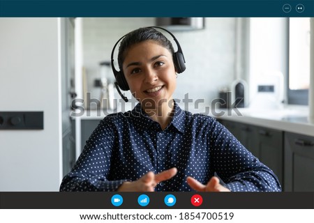 Head shot smiling beautiful young indian ethnicity businesswoman in headset with microphone enjoying pleasant video call distant conversation, sitting in kitchen, computer application screen view. Royalty-Free Stock Photo #1854700519