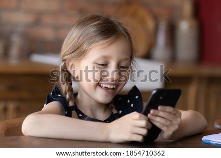 Kid and gadget. Happy laughing little child girl sitting at desk holding mobile phone in hands, making call to mother, chatting with friend, watching funny cartoon, posting selfie in social network