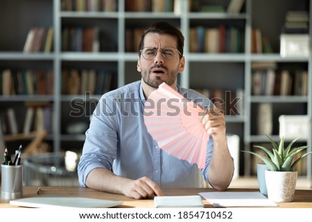 Man office manager sit at workplace desk hold pink handheld fan waving it to reduce unbearable hot weather suffers from high temperature indoor at summer workday in modern room without air conditioner Royalty-Free Stock Photo #1854710452