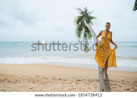 Happy young woman in yellow dress walking along the trunk of a palm tree on the beach