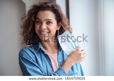 Happy Hispanic young woman takes off protective mask and looking at camera indoors while corona virus pandemic. Quarantine, and social distance concept.  Royalty-Free Stock Photo #1854759010