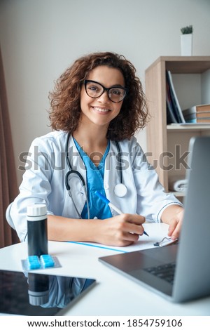 Vertical portrait of female doctor sitting on work desk and smiling at camera. Telemedicine, Medical online, e health concept. Doctor using laptop for work, video call video chat with colleagues. Royalty-Free Stock Photo #1854759106