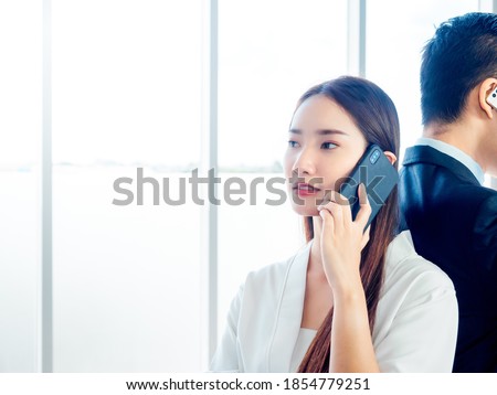Business Asian couple talking to mobile phone. Young beautiful confident woman calling with cell phone and standing back with businessman in suit on huge glass window background with copy space.
