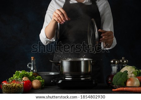 Professional chef salts boiling water in the pot on variety of ingredients background. Backstage of preparing appetizing food. Concept of cooking process. Frozen motion.