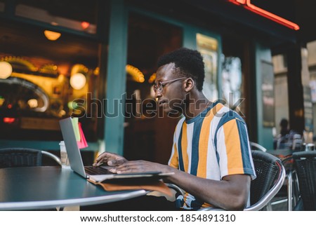 Skilled dark skinned student e learning via laptop application watching education webinar while sitting in street cafe and using public internet connection, African American freelancer reading text