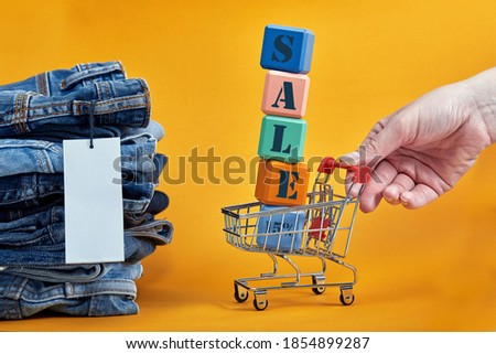 A stack of blue jeans with a white blank tag on a yellow background. Shopping trolley with multi-colored cubes. Sales word written on cubes. Sales consept