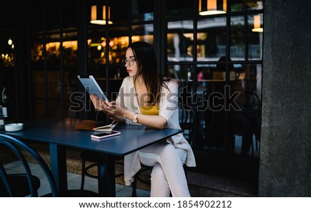 Millennial female blogger in spectacles holding modern digital tablet reading book online during break, beautiful female freelancer share publication and multimedia content via touch pad with 4g