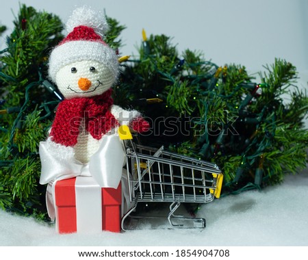 Empty grocery cart and toy snowman on red gift box in the snow with Christmas lights and Christmas tree background, place for text, black Friday discounts and sales, happy new year