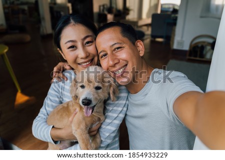 Young adult Asian couple holding a puppy taking a selfie from a phone with home interior in background. 30s mature man and woman with dog pet taking a family photo shots. - Happy group portrait