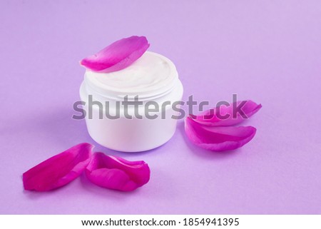 Jar with white delicate cream with peony extract and petals on pink background