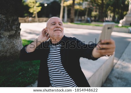 Elderly male blogger shooting influence vlog via modern cellphone application using roaming internet on vacations, aged senior clicking selfie smartphone pictures during daytime at city street