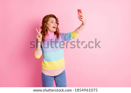 Photo portrait of cheerful girl saying hi over video chat making v-sign holding phone in one hand isolated on pastel pink colored background