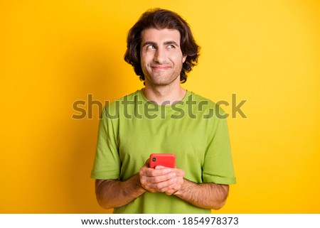 Photo of minded creative guy hold cellphone look up empty space wear green t-shirt isolated yellow color background