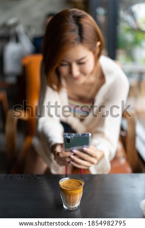 Blurred image of short-haired young Asian woman using cellphone taking a picture of coffee in a glass cup is placed on a wooden table in a coffee shop. Focus on smartphones and coffee in a glass cup.