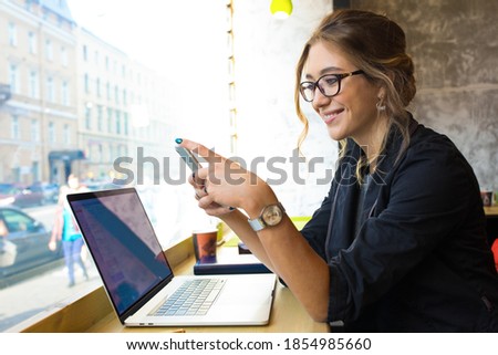 Happy smiling woman smart college student reading e-mail on mobile phone during online learning on laptop computer while sitting in coffee shop. Joyful female browsing wifi via cell telephone 