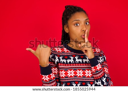 Young beautiful African American woman wearing Christmas sweater against red wall,  asking to be quiet with finger on lips pointing with hand to the side. Silence and secret concept.
