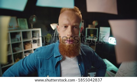 Portrait of joyful hipster guy looking at right note in office. Smiling business man picking up sticker from working board in dark office. Happy red head businessman finding good idea in workplace.