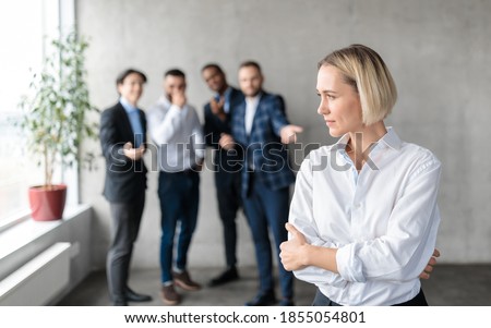 Sexism And Bullying At Work. Unhappy Victimized Business Lady Standing While Her Male Coworkers Whispering Behind Her Back In Modern Office. Workplace Discrimination Problem, Female Rights. Free Space Royalty-Free Stock Photo #1855054801
