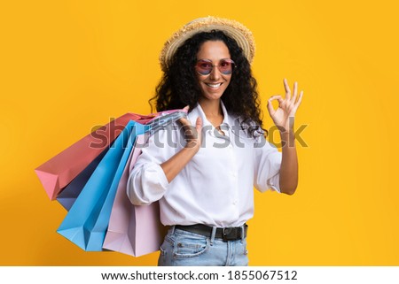 Big Sales. Young Brunette Woman Holding Shopping Bags And Showing Ok Sign, Enjoying Seasonal Discounts And Offers, Wearing Stylish Hat And Sunglasses, Posing Over Yellow Studio Background, Copy Space