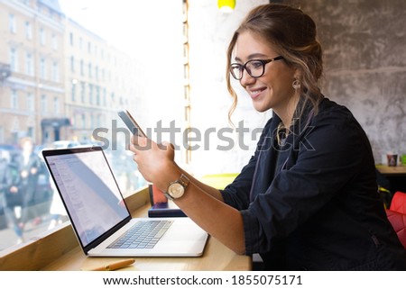 Happy smiling woman in glasses skilled business worker checking e-mail on mobile phone while sitting with laptop computer in coffee shop interior 
