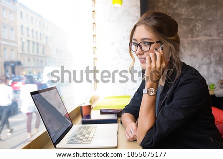 Female in spectacles smart college student talking via mobile phone during online learning on laptop computer while sitting in coffee shop during free time 