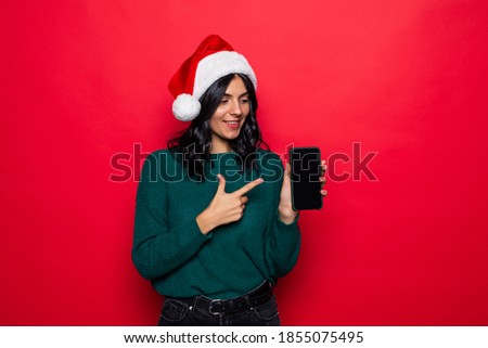 Smiling young woman in santa claus hat pointing finger at blank screen smartphone