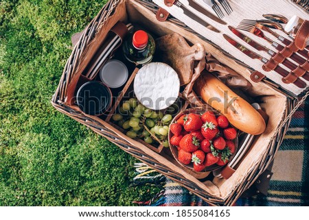 Picnic set with fruit, cheese, honey, strawberries, grapes, baguette, wine, wicker basket for picnic on plaid over green grass. Top view. Copy space. Summer picnic time, family lunch. Romantic picnic.