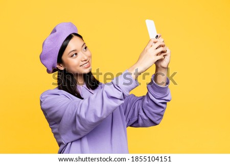 happy asian woman in beret taking selfie on smartphone isolated on yellow