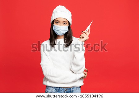 young asian woman in medical mask and hat holding digital thermometer isolated on red