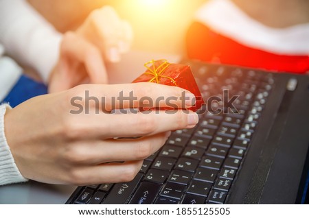 Woman holds a small red gift box in her hand against the background of a laptop with a Santa Claus hat, side view. Photo with illumination. The concept of Christmas, New year, holiday.