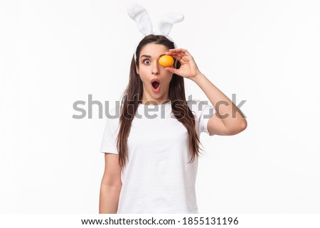 Waist-up portrait of amazed and surprised young woman seeing something awesome, hold colored egg over eye, staring shocked camera, gasping impressed, wear rabbit ears, celebrate Easter day