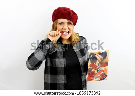 Beautiful female painter with cap and plaid shirt on isolated white background, she holds a palette of colors in one hand and in the other she holds the brush between her teeth in a grimace of anger. 