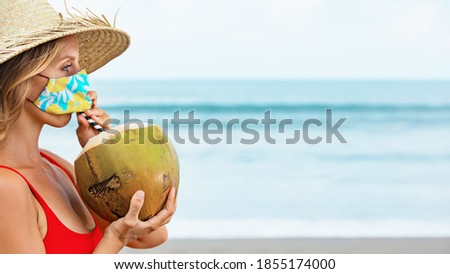 Funny portrait of woman in straw hat drinking young coconut on tropical sea beach. New rules to wear cloth face covering mask at public places due coronavirus COVID 19. Family holiday, summer travel