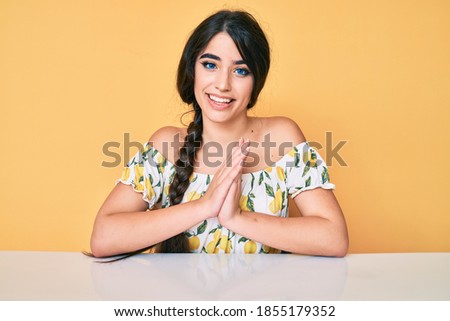 Brunette teenager girl wearing casual clothes sitting on the table praying with hands together asking for forgiveness smiling confident. 