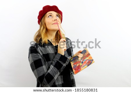 Beautiful woman painter in a cap and plaid shirt holds a palette of colors in one hand and in the other holds the brush on the tip of her nose looking up for inspiration. Creative crisis concept