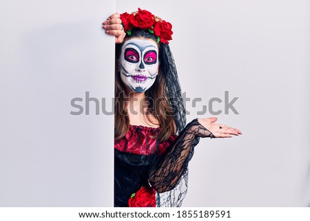 Young woman wearing day of the dead custome holding blank empty banner smiling cheerful presenting and pointing with palm of hand looking at the camera. 