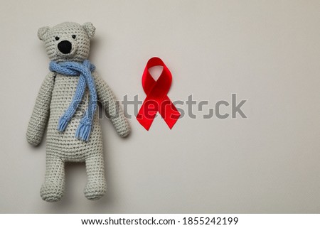 Cute knitted toy bear and red ribbon on beige background, flat lay with space for text. AIDS disease awareness