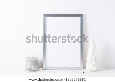 Photo or poster mockup with a silver frame, small decorative ceramic christmas tree  and candle on white table near a light wall. Home  holiday interior